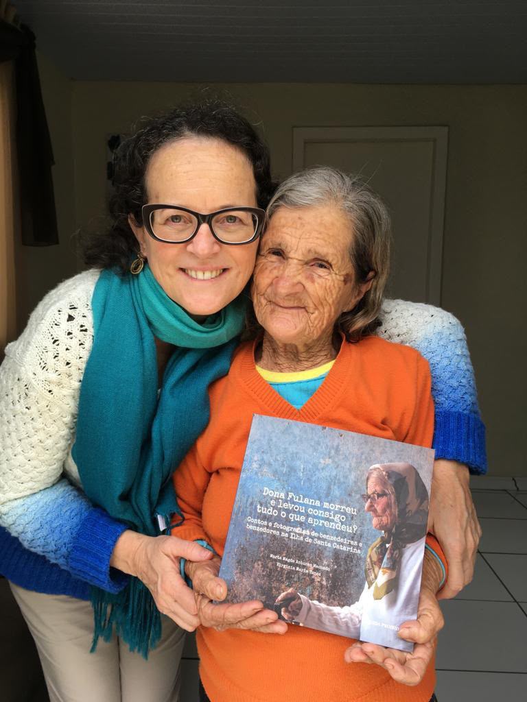Marta Magda abraçada com uma das benzedeiras entrevistadas, segurando o livro Dona Fulana morreu e levou consigo tudo o que aprendeu?